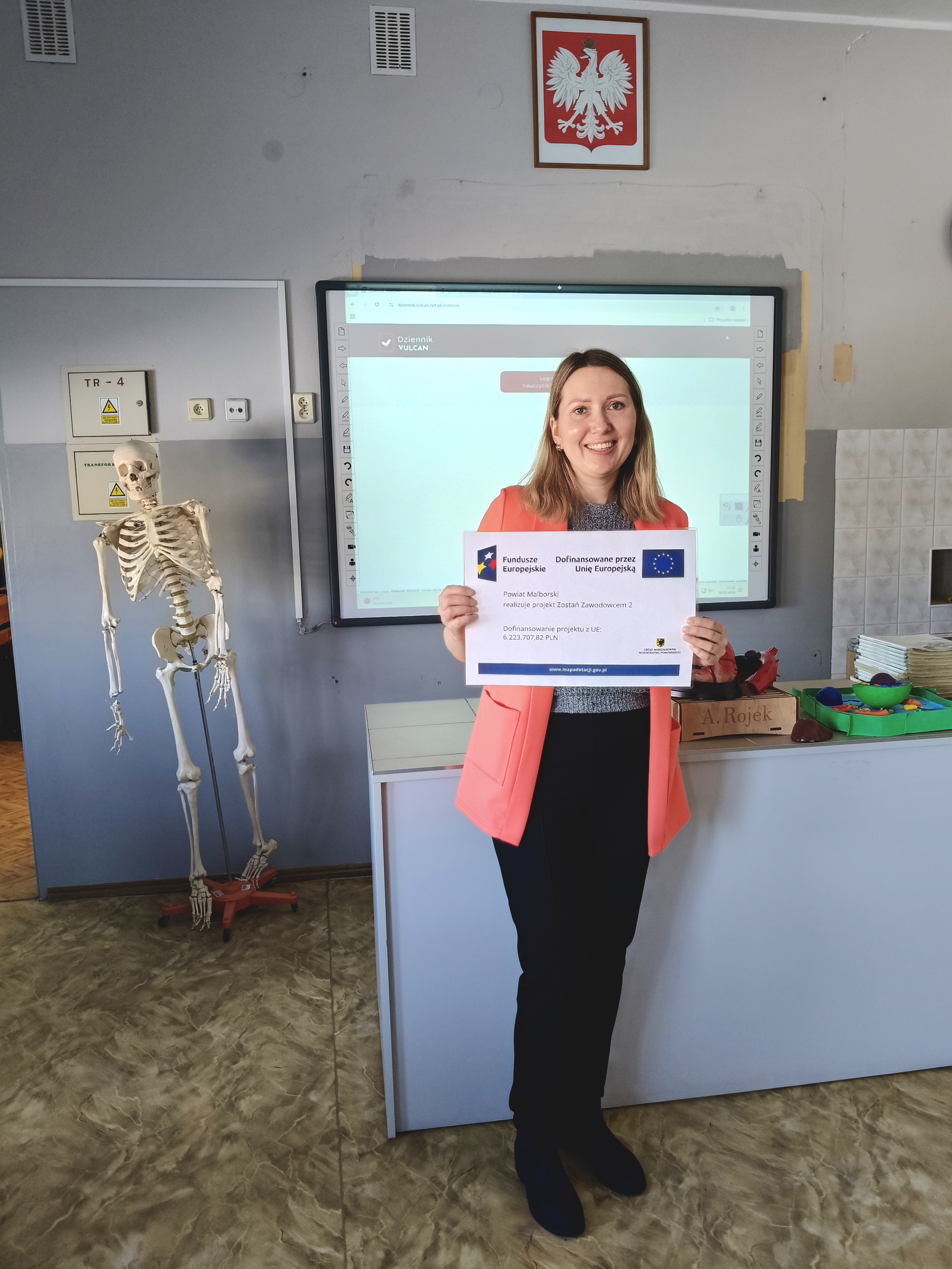 Zdjęcie przedstawia Koordynatorkę Powiatową na tle klasy, trzymającą plakat informujący o realizacji projektu pn. Zostań Zawodowcem 2 